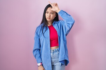 Young asian woman standing over pink background making fun of people with fingers on forehead doing...