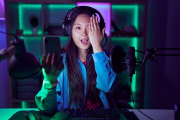 Young asian woman playing video games with smartphone covering one eye with hand, confident smile on face and surprise emotion.