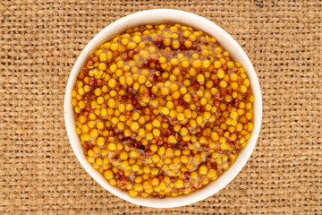French mustard in a white saucer on a jute cloth, macro, top view.