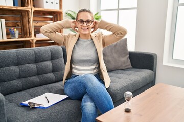 Obraz premium Young woman working at consultation office relaxing and stretching, arms and hands behind head and neck smiling happy