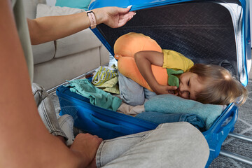 Family mother and child are having fun, the child is hiding in a suitcase, packing things and travel pillow for a trip