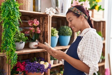 Young beautiful hispanic woman florist holding plant on shelving reading notebook at florist