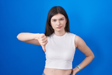 Young caucasian woman standing over blue background looking unhappy and angry showing rejection and negative with thumbs down gesture. bad expression.