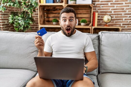Handsome hispanic man doing online shopping holding credit card scared and amazed with open mouth for surprise, disbelief face