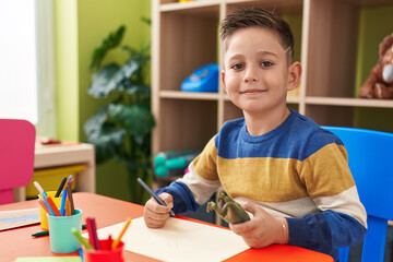 Adorable hispanic boy student sitting on table drawing on paper at kindergarten