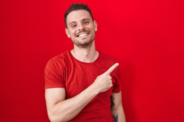Young hispanic man standing over red background cheerful with a smile on face pointing with hand and finger up to the side with happy and natural expression