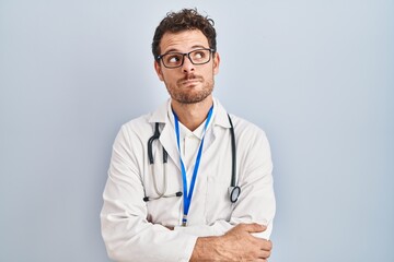 Young hispanic man wearing doctor uniform and stethoscope smiling looking to the side and staring away thinking.