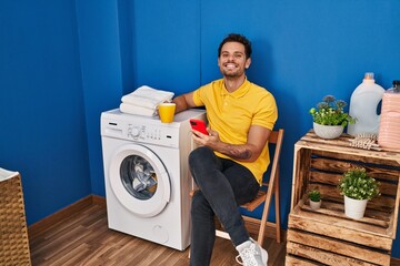Young hispanic man drinking coffee using smartphone wating for washing machine at laundry room