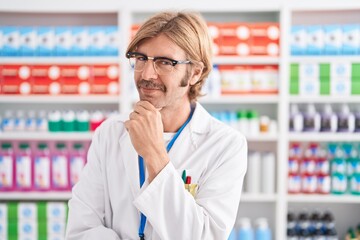 Caucasian man with mustache working at pharmacy drugstore looking confident at the camera smiling with crossed arms and hand raised on chin. thinking positive.