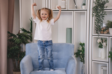 Fototapeta na wymiar Little blonde caucasian girl with ponytails rises hands with clenched fists looks at camera happily waits for present on holiday. Healthy Italian child smiles, celebrating. Childhood, domestic leisure