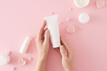 Women's cosmetics concept. First person top view photo of female hands holding tube cream, heart shaped candles, cream jars and heart baubles on pastel pink background. Mother's day cosmetic idea.