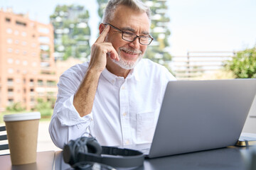 Happy older senior business man using laptop elearning sitting outdoor. 60 years old professional businessman working online on computer, watching webinar, learning or having remote video call.