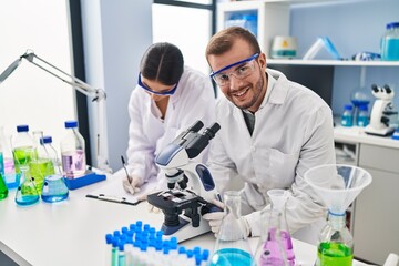 Man and woman scientist partners working using microscope and writing on clipboard at laboratory