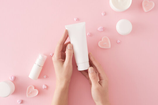 Women's cosmetics concept. First person top view photo of female hands holding tube cream, heart shaped candles, cream jars and heart baubles on pastel pink background. Mother's day cosmetic idea.