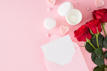 Mother's day concept. Flat lay photo of cream jars, red roses, heart shaped candles and envelope with letter on pastel pink background with copy space. Women's day cosmetic idea.