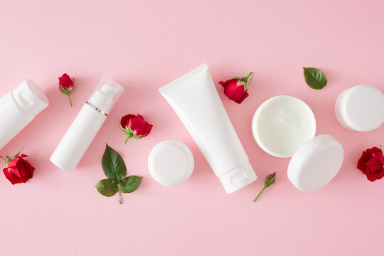 Women skin care concept. Flat lay photo of cream jars, white tubes without label, red roses and leaves on pastel pink background. Mother's day cosmetic idea.