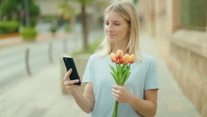 Young blonde woman using smartphone holding bouquet of flowers at street