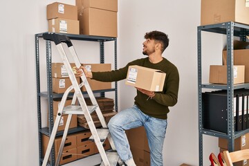 Young arab man ecommerce business worker holding package standing on ladder at office