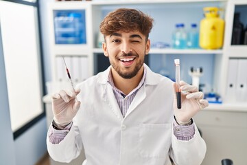 Arab man with beard working at scientist laboratory holding blood sample winking looking at the...