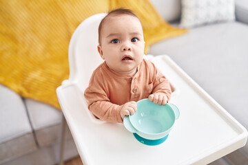 Adorable hispanic toddler holding bowl sitting on highchair at home
