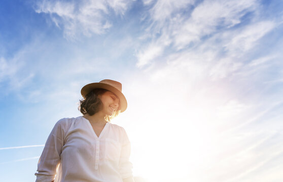 Young dreamy beautiful woman in a hat and white shirt standing against a blue sky with clouds
