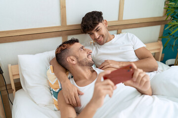 Two hispanic men couple lying on bed watching video on smartphone at bedroom