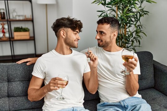 Two hispanic men couple toasting with glass of wine holding key of new house at home