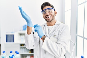 Young hispanic man working at scientist laboratory putting gloves on smiling and laughing hard out loud because funny crazy joke.
