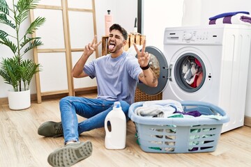 Young hispanic man putting dirty laundry into washing machine smiling with tongue out showing fingers of both hands doing victory sign. number two.