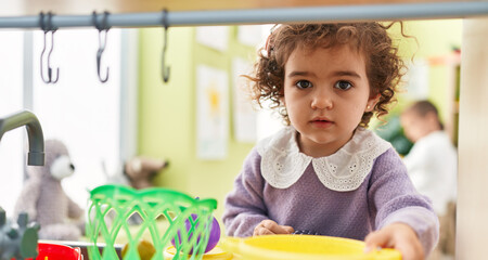 Adorable hispanic girl playing with play kitchen standing at kindergarten