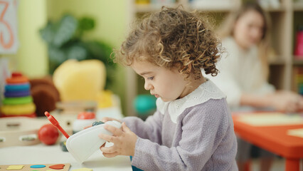 Adorable hispanic girl holding gift sitting on floor by christmas tree with unhappy expression at kindergarten