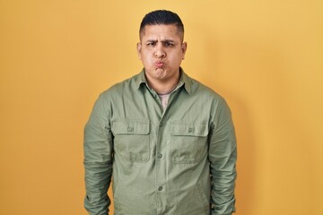 Fototapeta premium Hispanic young man standing over yellow background puffing cheeks with funny face. mouth inflated with air, crazy expression.