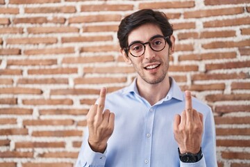 Young hispanic man standing over brick wall background showing middle finger doing fuck you bad...
