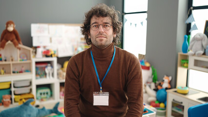 Young hispanic man preschool teacher standing with relaxed expression at kindergarten