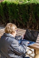 Senior woman sitting outdoors on a bench using a laptop, on a sunny winter day.