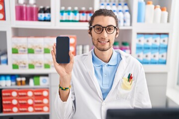 Young hispanic man working at pharmacy drugstore showing smartphone screen looking positive and happy standing and smiling with a confident smile showing teeth