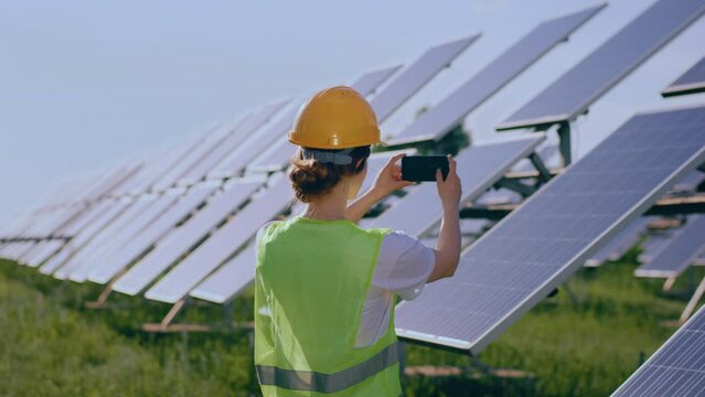 Taking video at photovoltaic solar farm ecological engineer woman with safety equipment taking pictures of solar panels with the smartphone