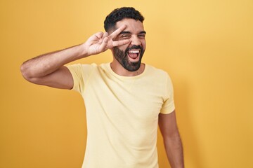 Hispanic man with beard standing over yellow background doing peace symbol with fingers over face, smiling cheerful showing victory