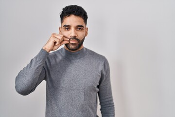 Hispanic man with beard standing over white background mouth and lips shut as zip with fingers. secret and silent, taboo talking