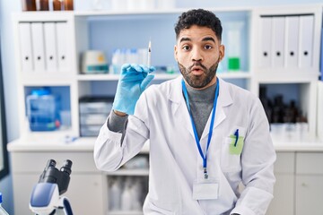 Hispanic man with beard working at scientist laboratory holding syringe scared and amazed with open...