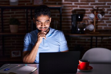 Fototapeta na wymiar Hispanic man with beard using laptop at night looking confident at the camera smiling with crossed arms and hand raised on chin. thinking positive.
