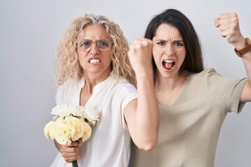 Mother and daughter holding bouquet of white flowers angry and mad raising fist frustrated and furious while shouting with anger. rage and aggressive concept.