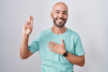 Middle age bald man standing over white background smiling swearing with hand on chest and fingers...