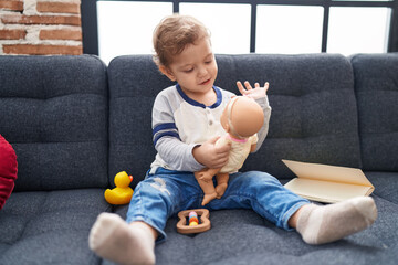 Adorable caucasian boy playing with baby doll sitting on sofa at home
