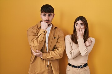 Young hispanic couple standing over yellow background looking stressed and nervous with hands on mouth biting nails. anxiety problem.