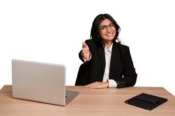 Young indian woman in a table with a laptop and tablet isolated stretching hand at camera in greeting gesture.