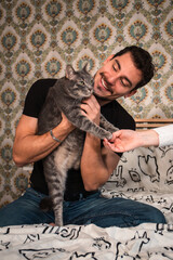 The young man is playing with his cat on the bed. Beautiful man is stroking the fluffy gray cat. - 561593979