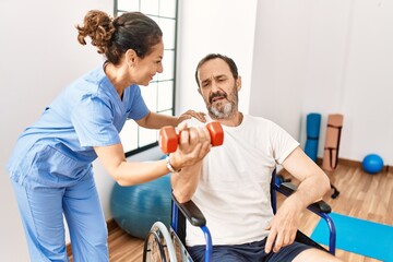 Middle age man and woman having rehab session using dumbbell sitting on wheelchair at physiotherapy clinic