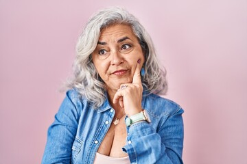 Middle age woman with grey hair standing over pink background thinking worried about a question, concerned and nervous with hand on chin