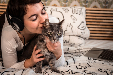 Beautiful woman plays with the cat on the bed. The young woman listens to music on headphones while she is petting her cat. - 561596546
